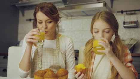 Portrait-shot-of-the-attractive-mother-and-her-pretty-teen-daughter-sitting-in-kitchen,-eating-muffins,-drinking-juice-and-hugging.-Indoor